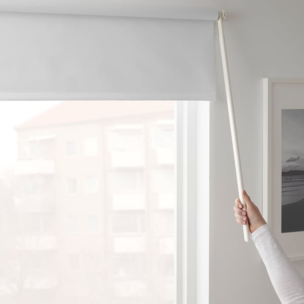 Hand holds wand to control white roller blind, adjusting light and privacy in room.