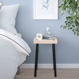 Small bedside table: IKEA FRIDNÄS. Light wood top, black legs. Holds clock, book, glass. Next to bed, blue wall, artwork.