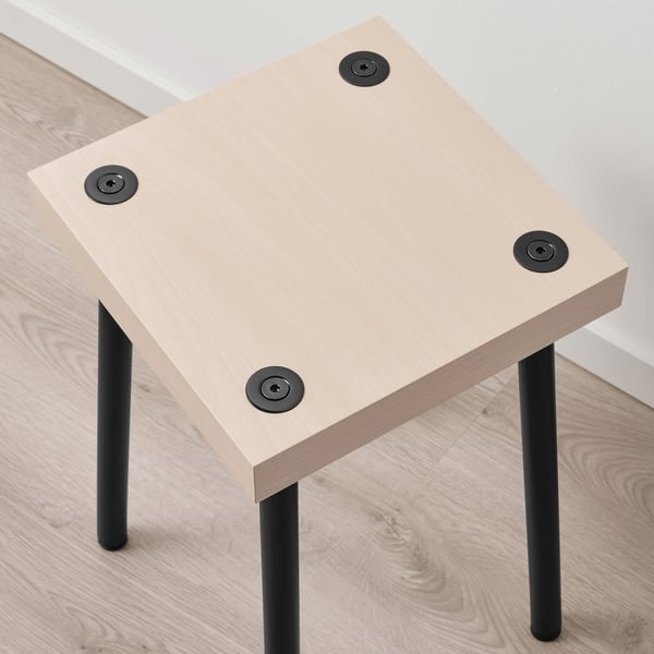 Square side table with light-colored wooden top; four black metal legs with adjustable feet, sitting on a wooden floor beside a white wall.