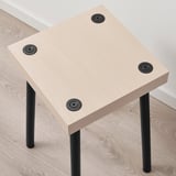 Square side table with light-colored wooden top; four black metal legs with adjustable feet, sitting on a wooden floor beside a white wall.