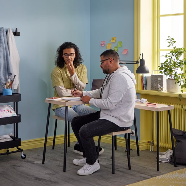 Two people sit at FRIDNÄS nesting tables, one writing and the other thinking. Light wood tables, black metal legs, blue wall, yellow accents.