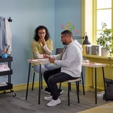 Two people sit at FRIDNÄS nesting tables, one writing and the other thinking. Light wood tables, black metal legs, blue wall, yellow accents.