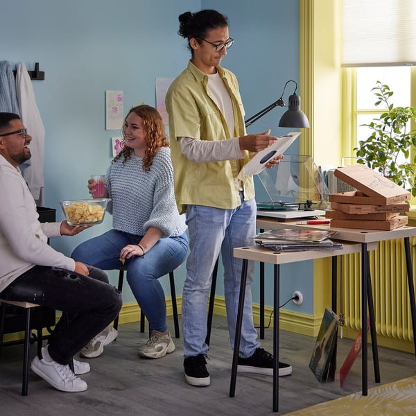 Three people in a cozy blue room. One holds a vinyl, another chips. Two wooden tables with records and food. A lamp lights the scene.