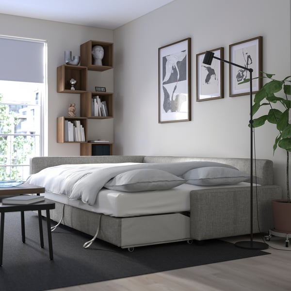 Modern bedroom with gray FRIHETEN sofa-bed, white bedding, and wooden bookshelf. Natural light from window, adjacent plant, and wall art.
