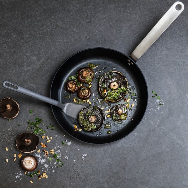 Top view of black non-stick frying pan with mushrooms, herbs, and oil. Silver spatula beside. Gray background with scattered mushrooms and seasoning.