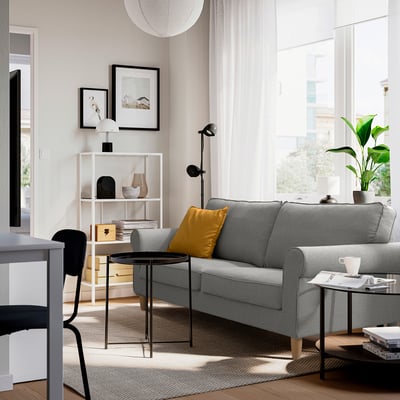 Modern living room with grey GÄLLTOFTA sofa, yellow pillow, black side table, white curtains, large window, and plants.