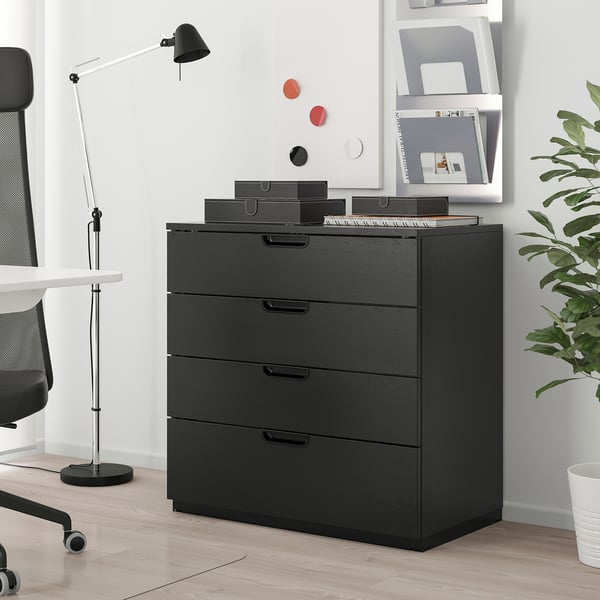 GALANT ikea chest: black 4-drawer unit beside desk, chair, lamp. Top has boxes, papers. Background: whiteboard, bookshelf.
