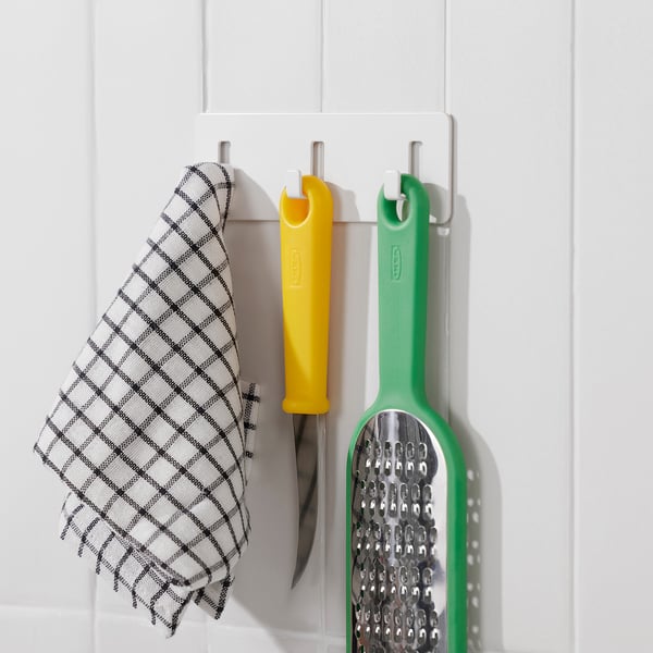 Three hooks on a white wall hold a checkered cloth, a yellow knife, and a green grater. Organizes kitchen tools efficiently.