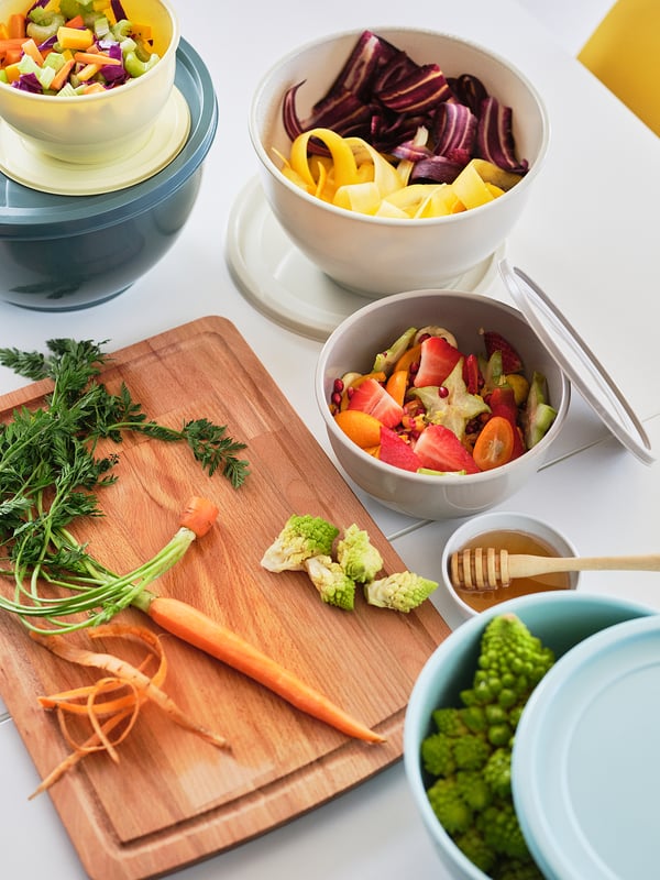 Colorful vegetables prepared on wooden cutting board, multiple bowls, one with honey, all arranged on white surface.