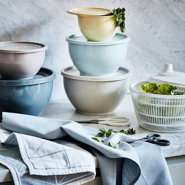 Stack of colorful bowls in blue, green, and cream, with lids, on a marble countertop, surrounded by kitchen utensils.