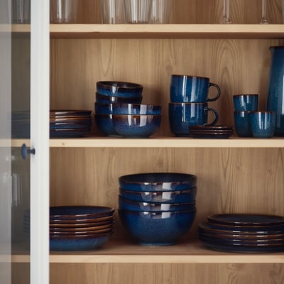 Shelf with blue GLADELIG stoneware mugs and bowls, handcrafted look, stacked neatly, golden-brown details.