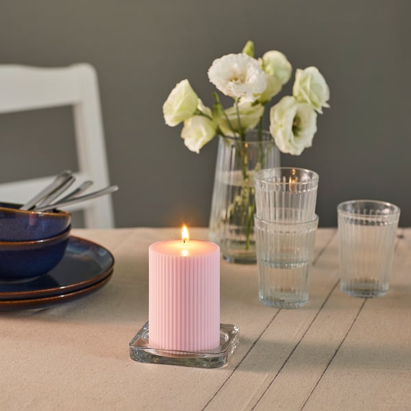 Pink GLASBJÖRK candle in glass holder with flowers, glasses on table, cosy atmosphere.