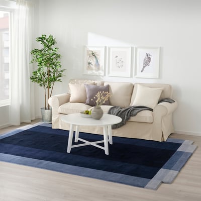 Living room with GNETGRÄS rug, blue shades, wool. Sofa, coffee table, and plant beside. Rug large, fits under furniture.