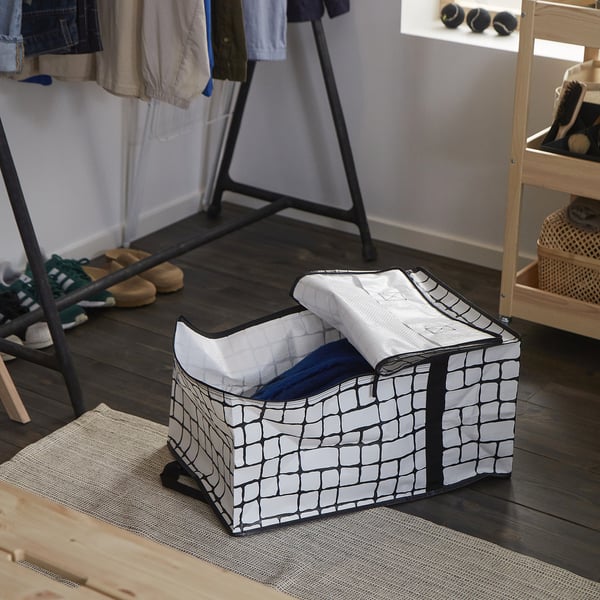 A GÖRSNYGG storage bag on wooden floor, gray-black grid pattern, partially filled with clothes, near shoe rack.