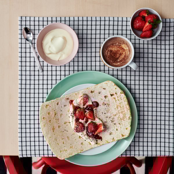 A meal setup on a checkered tablecloth featuring a folded crepe with berries, a cup of coffee, a bowl of cream, and a bowl of strawberries.