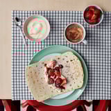 A meal setup on a checkered tablecloth featuring a folded crepe with berries, a cup of coffee, a bowl of cream, and a bowl of strawberries.