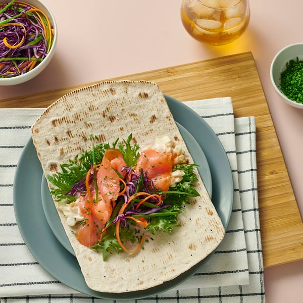 A flatbread wrap with salmon, greens, and sauce on a green plate. Surrounded by bowls of salad and herbs.