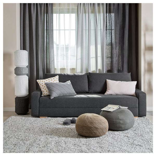 Modern gray sofa-bed with colorful pillows, round poufs, and tall white lamp.