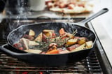 A black cast iron skillet on a grill cooks vegetables with herbs, emitting steam. A pizza bakes on a tray above. Ideal for outdoor bbq parties.