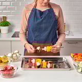 Woman grilling kebabs using GRILLTIDER tray with various vegetables.