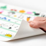 Hand peels back corner of colorful GRÖNFINK place mat, revealing grip pattern, designed for kids, protecting tables from spills.