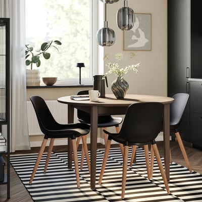 Modern dining set with black chairs, round light wood table, striped rug, hanging lamps, and large window with plants.