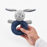A hand holds a grey and blue polka dot bunny rattle toy, designed for baby to grip, shake, and chew safely.