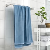 GULVIAL blue towel hangs on bathroom rail, next to white shelves holding folded towels and a green plant.