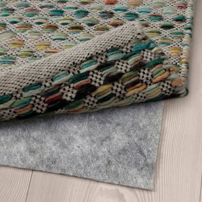 Multicolored woven rug on a wooden floor with a gray non-slip mat underneath ensuring stability.