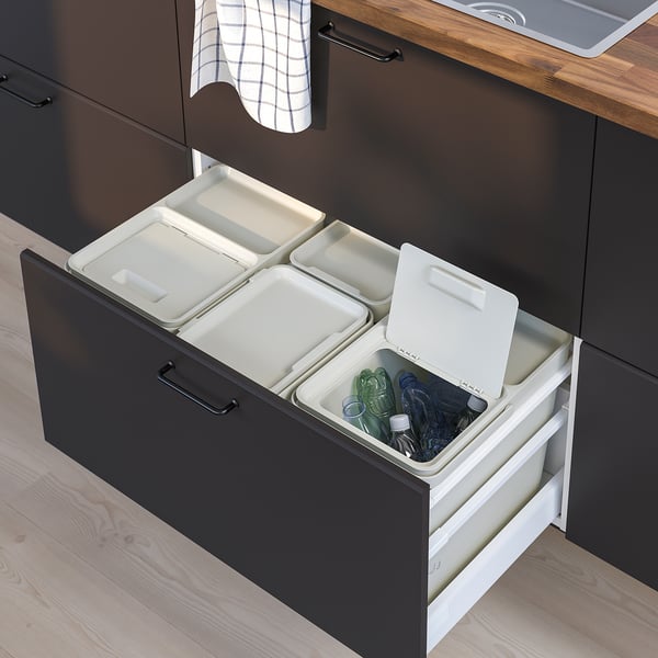 Modern kitchen drawer with HÅLLBAR waste sorting bins in white, black, wood floor, lids open, various items inside.