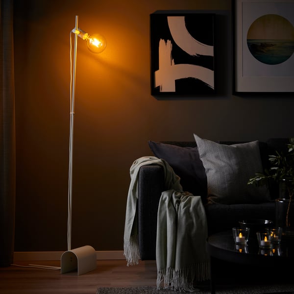 HÅRSLINGA floor lamp with warm light, positioned beside a couch with blankets & pillows, against a painting-adorned wall. Tall, slim design.