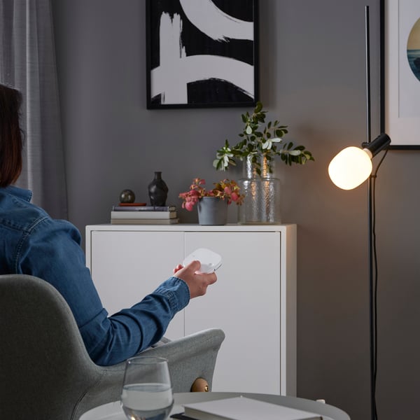 Person sitting in chair, holding remote, controlling smart lamp with warm light, beside cabinet with plants and decor.
