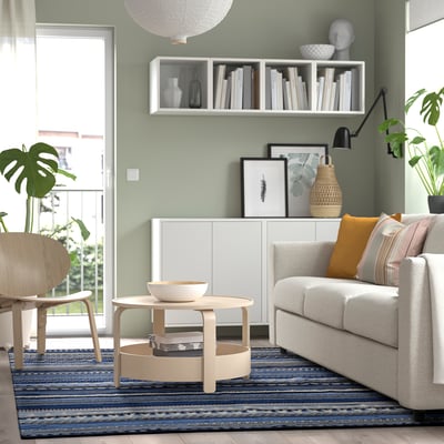Modern living room with green walls, blue rug, white sofa, wooden coffee table, sideboard, plants, lamp, and balcony door.