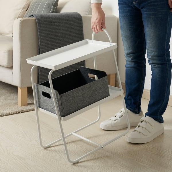 White side table with shelves and bin, next to beige chair. Person stands nearby, showing mobility.