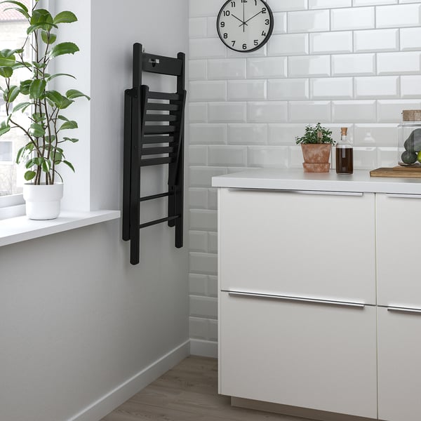 Small modern kitchen with white cabinets, a hanging black foldable chair, and a clock on white tiles. A plant on the windowsill adds greenery.