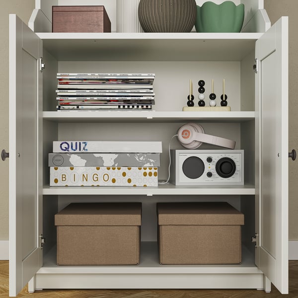 White HAUGA storage cabinet with shelves, containing books, games, headphones, and fabric boxes; ideal for organizing and displaying items.