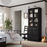 Modern bedroom with dark HAVSTA drawers & glass cabinet, white walls, metal bed, plant, chair, rug.