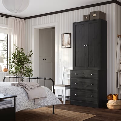 Bedroom with dark HAVSTA cabinet, white chair, black bed, rug, and plant.