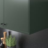 Green kitchen cabinet with sleek handle, hiding underlighting. Plant and pepper mill nearby.