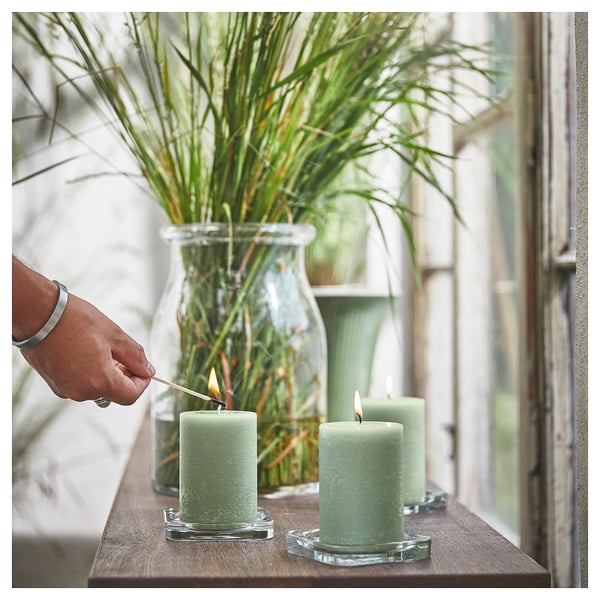 A person lights green HEDERSAM candles on a table beside a glass vase with grassy plants.