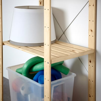 A wooden shelf unit with a white lamp on top and a clear bin of pool noodles stored on a middle shelf.