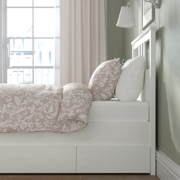 Bed with ornate bedding, large pillows, and white frame. Storage boxes underneath. Light through sheer curtains.