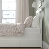 Bed with ornate bedding, large pillows, and white frame. Storage boxes underneath. Light through sheer curtains.