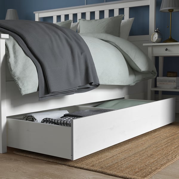 Under-bed drawer for storage, with folded blankets inside. Bed has gray and white bedding, a nightstand with a lamp nearby.