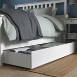 Under-bed drawer for storage, with folded blankets inside. Bed has gray and white bedding, a nightstand with a lamp nearby.