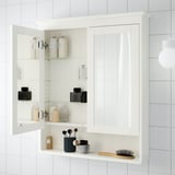 White wall-mounted bathroom mirror cabinet with adjustable glass shelves, storing toiletries and makeup items.