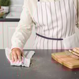 Person wearing apron cleans kitchen counter with tea towel, beside cutting board with bread slices.