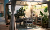 Cozy outdoor dining setup with grey HIMMELSÖ gazebo, wood table, black chairs, plants, and string lights.