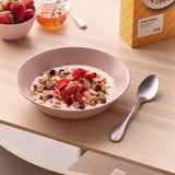 A bowl of granola with yogurt, strawberries, and other berries is shown on a table with a spoon.