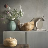 Styled natural scene: green vase, woven baskets, driftwood circle on grey shelf.